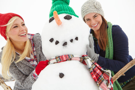 Women, friends and snowman on vacation, laugh or bonding for fun, games or excited in winter. People, happy and cold weather at holiday resort, playful or sweater for getaway with sculpture in natureの写真素材