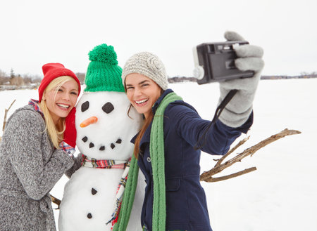 Women, friends and selfie with snowman, camera and bonding with hug, happy and winter holiday. People, memory and photography with cold weather, vacation and embrace sculpture with laugh in natureの写真素材