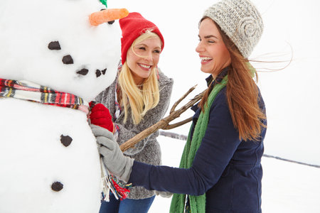 Women, friends and building snowman, happy and outdoor for bonding with fun, games or winter holiday. People, smile and cold weather with break, vacation or playful with sweater with branch in natureの写真素材