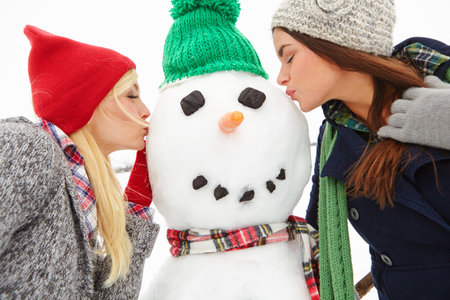 Women, friends and kiss with snowman, holiday or bonding for fun, memory and care in winter. People, happy and cold weather with love, vacation and building ice sculpture with connection in natureの写真素材