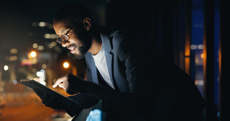 Black man, tablet and night at office, balcony and smile with typing, review and check notification on break. African person, writer and happy with tech, app or overtime for job at creative agencyの写真素材