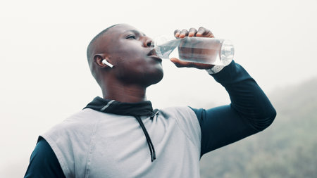 Fitness, black man outdoor and drink water with earbuds for music playlist, workout break or below. Person, beverage and audio tech in nature with streaming service, album and hydration for exercise.の写真素材