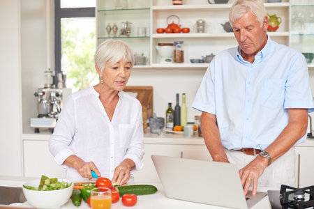 Old couple, chop vegetable or talk in kitchen with laptop for healthy diet, meal prep or cooking website. Married, elderly people or cut ingredients in home with computer, food recipe or instructionsの写真素材