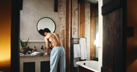 Back, basin and doorway with shirtless man in bathroom of hotel room for hygiene or morning routine. Entrance, start and wake up with person at sink for accommodation, break or hospitality as guestの写真素材
