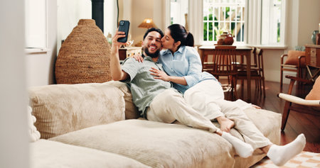Happy couple, selfie and hug on couch, home and web post with cheek kiss, love or relax in lounge. Woman, man and smile for photography on sofa, profile picture or embrace on social media at houseの写真素材