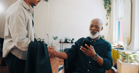 Senior man, barber and handshake with phone for hairstyle, option or choice in beauty salon. Elderly, male person or customer shaking hands with hairdresser on mobile smartphone for haircut serviceの写真素材