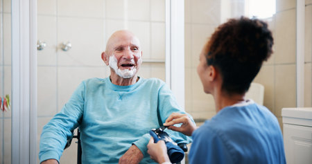 Senior man, nurse and shaving in bathroom with wheelchair, happy and talk with grooming in home. People, caregiver and elderly person with disability, razor or cream for facial hair removal at houseの写真素材