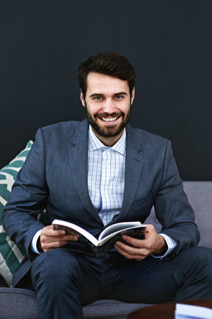Smile, book and portrait of businessman reading on sofa in waiting area for corporate knowledge at office. Happy, career and male financial advisor enjoying finance literature on couch in workplace.の写真素材