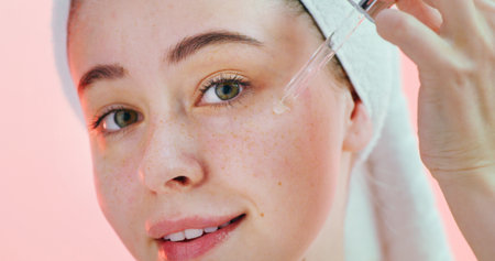 Beauty, face and girl with serum in studio for hyaluronic acid, hydration and healthy skincare routine. Gen z, person and retinol oil application on pink background for vitamin c treatment and glowの写真素材