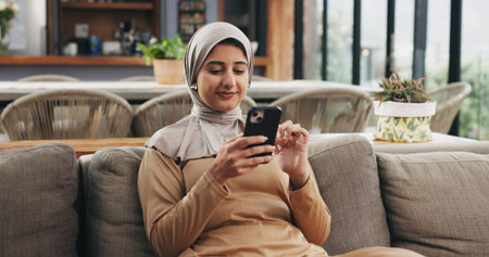 Muslim woman, phone and happy with texting on sofa in home, scroll or check notification in lounge....