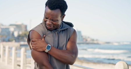 Black man, shoulder pain and fitness with injury on beach promenade on break with sore muscle in summer. Person, runner and fatigue with joint strain, exercise or rest with space on seaside boardwalkの写真素材