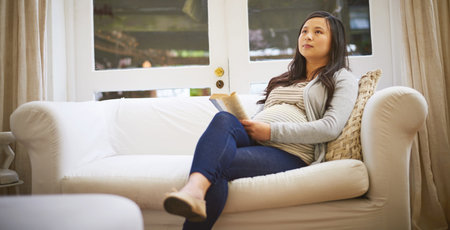 Thinking, book and research with pregnant woman on sofa for parenting advice, study baby growth and...