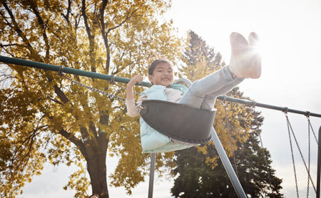 Smile, portrait or kid at playground on swing, growth or adolescent fun in winter holiday. Low angle, playful or Asian girl at park with flare, outdoor activity or child development on weekend break.の写真素材