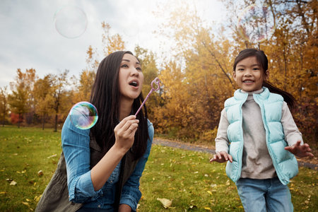 Asian, child and mother blowing with bubbles in park for playful childhood or outdoor bonding. Mom,...