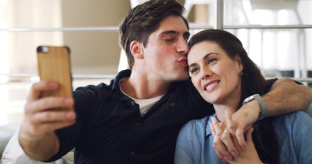Happy couple, selfie and kiss on sofa in home with bonding, connection or care with web post. Man, woman and smile with partner, photography or hug with love for social media on couch at apartmentの写真素材