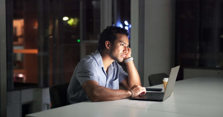 Bored, laptop and night with business man in office for planning fatigue or research deadline. Computer, reading and tired with exhausted employee at desk in dark workplace for overtime burnoutの写真素材