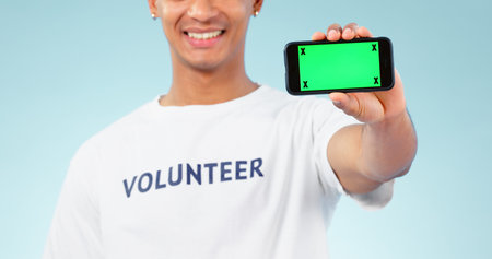 Hand, phone and green screen with a volunteer man in studio on blue background for marketing or charity. Smile, space or advertising and a person showing chromakey on a display with tracking markers