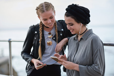 Phone, friends and women outdoor for music, social media and sharing playlist by promenade. Smile, mobile and girls hangout with audio app, earphones and show podcast info on internet by ocean