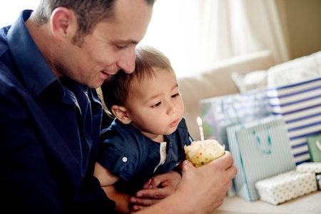 Dad, child and cupcake with candle for blowing, childhood and bonding on sofa in home. Father, son and wish for celebration, love and fun in living room of house for event together with presents