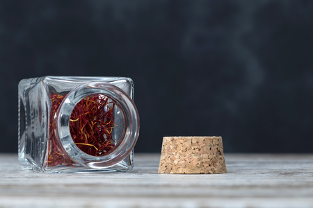 saffron threads inside a small glass jar opened on a wooden table close to the corkの写真素材
