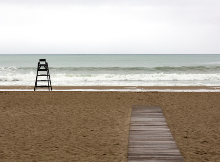 the beach during a cloudy day with a beach watchman chair alone on the sand and a wooden pathwayの写真素材