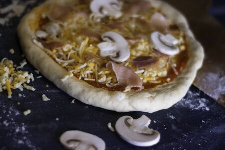 Homemade pizza dough with cheese, mushrooms, oregano, tomato, ham and flour over dark background with white and orange lights.の写真素材