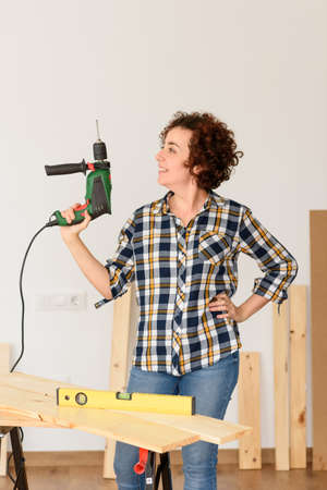 Caucasian woman with curly hair holds a drill in her hands, ready to make home improvements. She is standing in front of a workbench. She wears a plaid shirt and jeans. White background.の写真素材