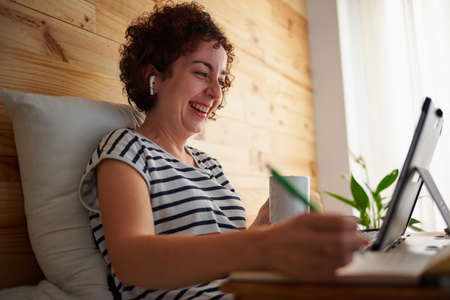 A curly-haired woman drinks coffee while working from her bedroom. The room has a nice wooden wall and it is daytime. New technologies allow you to work comfortably. She uses a folding table.の写真素材