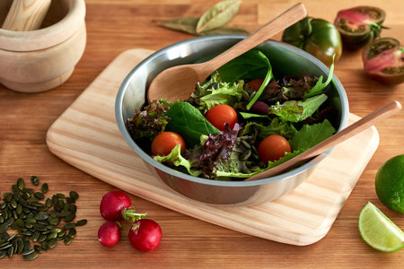 Salad of green sprouts, lettuce, spinach, cherry tomato, seeds, radish, lime and bay leaves, served in a metal bowl on a wooden cutting board on a restaurant kitchen table next to a wooden mortar.の写真素材