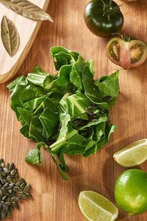 Spinach leaves on a wooden kitchen table. There is a cutting board with bay leaves, tomatoes, lime,...