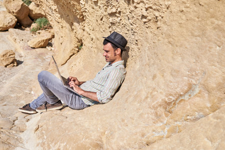 A man with a hat using the keyboard of his laptop in a tourist beach area with his camera and his...