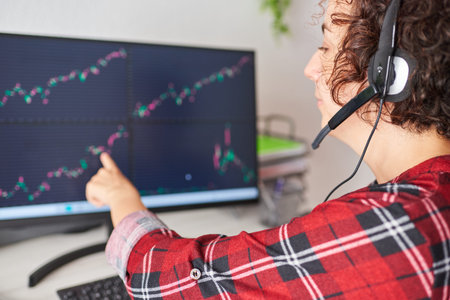 Young businesswoman points to computer screen with stock market graphs while wearing headset