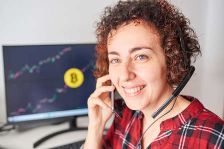 Female trader operates with bitcoins from her standing desk while talking on the mobile phone and wearing a headsetの写真素材