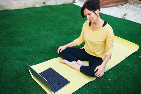 A woman takes a yoga class from her laptop outdoorsの写真素材