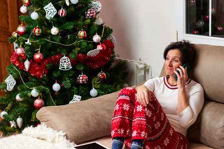 Woman with sad face sitting on the couch by the christmas tree while talking on the phoneの写真素材