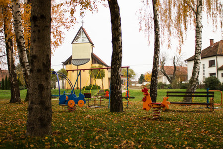 Colorful playground in park covered with yellow fallen autumn leaves and yellow church from behindの写真素材