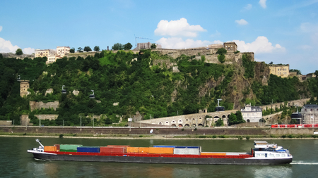 A colorful, long, loaded container boat sails on the Rhine River and the green hills above it, while a red, electric train passes in the meantime people are driving in the funiculars above themのeditorial素材