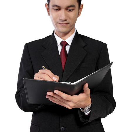 Low angle shot of young Asian man in formal suit writing on a notebook, isolated on white backgroundの写真素材