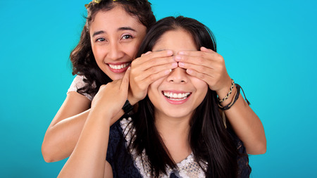 Two smiling Asian female friends play guess who game. One girl covering her friend's eyes from behind, over blue backgroundの写真素材