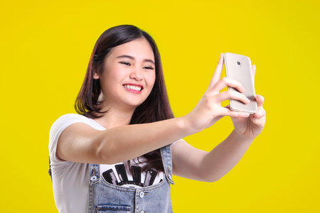 Cute smiling Asian teenage girl taking self shot photo with her phone camera, on bright yellow...