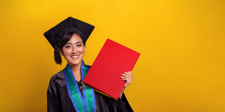 Young attractive Asian female student on her graduation day, holding a red colored certificate with happy face, over bright yellow studio backgroundの写真素材