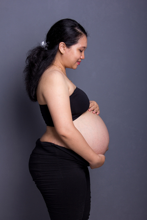 Side view portrait of pregnant lady in black outfits, watching over her unborn baby, standing on grey backgroundの写真素材