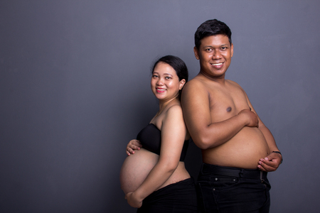 Pregnant mom and dad holding their big tummies, smiling and posing at camera, studio shot on grey backgroundの写真素材