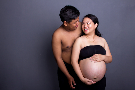 Lovely Asian ethnic couple during maternity. Pregnant lady and her husband looking at each other. Over grey studio backgroundの写真素材