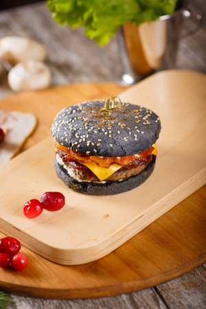 Close up Modern black Burger with beef meat, bacon, cheese, lettuce and vegetables garnished with fried potato wedges and barbecue sauce on the wooden standの写真素材