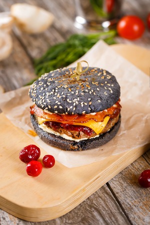Close up Modern black Burger with beef meat, bacon, cheese, lettuce and vegetables garnished with fried potato wedges and barbecue sauce on the wooden standの写真素材