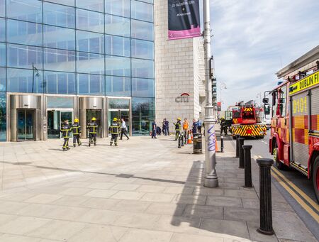 Firefighters and two fire engines outside office building. Dublin, Ireland - April 21, 2016: Two fire engines and a group of firefighters from Dublin Fire Brigade outside a building.  Firefighters talking. People passing by.のeditorial素材