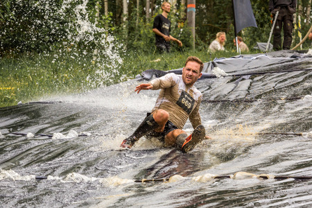 Stockholm, Sweden - June 03, 2017: Front view of one caucasian man riding a muddy water slide down hill at an obstacle course to complete the annual event Toughest Stockholm.のeditorial素材