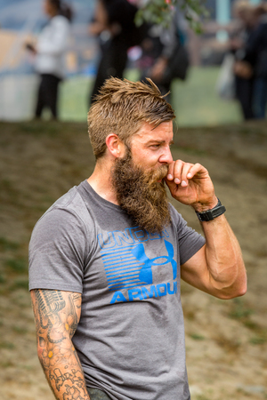 Stockholm, Sweden - June 03, 2017: Profile view of athletic sweaty young man with full beard resting after reaching the finish line in the event Toughest Stockholm. Blurred people in the background.のeditorial素材