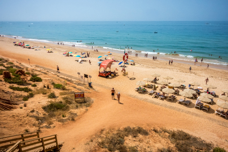Aerial view of beach. Albufeira, Portugal - July 16, 2017: Landscape view from above of many people on public beach at the Algarve coast sunbathing and walking along the waterline. Stairs to the beach and sun parasols to get some shade.のeditorial素材